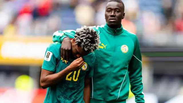 Pape Gueye, vêtu d'un survêtement vert du Sénégal, regarde la caméra tout en consolant Ibrahim Mbaye, un bras autour de l'épaule de son coéquipier qui baisse les yeux et porte sa main droite à son visage. Les deux joueurs sont visibles jusqu'à la taille, et une tribune de stade de football se devine en arrière-plan, légèrement floue.