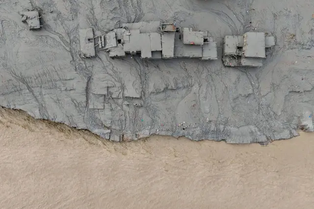 Un río marrón pasa al lado de una serie de construcciones cubiertas de barro debido a una avalancha reciente. La foto es tomada desde arriba. 