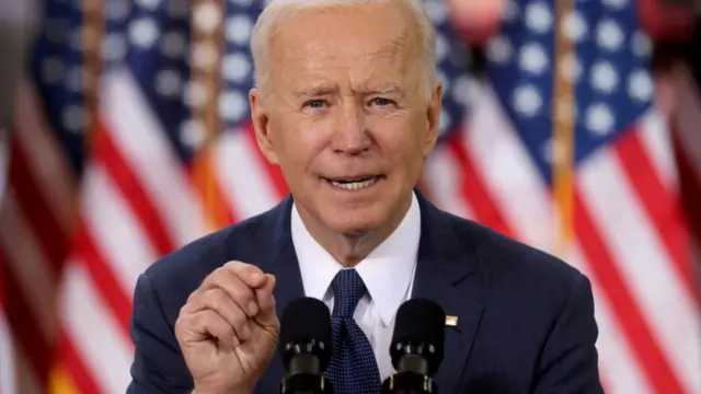 Biden gesticula enquanto fala no microfone, com bandeiras dos EUA atrás