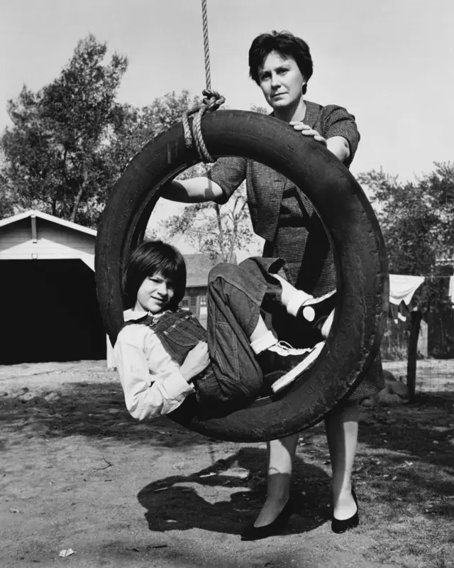 Девочка с короткой стрижкой и в комбинезоне сидит на импровизированных качелях из шины, висящей на веревке. Ее качает женщина с короткой стрижкой, одетая в платье. 