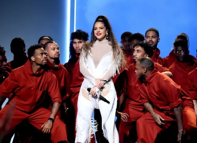 Rosalía en el escenario, vestida de blanco con un micrófono y acompañada de un grupo de bailarines vestidos de rojo