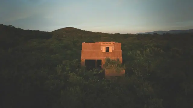 Edificio de concreto rojizo aislado, emergiendo entre vegetación densa en un paisaje montañoso al atardecer.