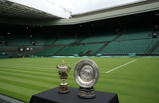 Widok na puchar w grze pojedynczej mężczyzn i puchar w grze pojedynczej kobiet na stoliku przykrytym czarnym obrusem przed turniejem Wimbledon, w tle kort tenisowy