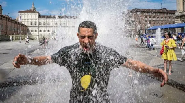 Homem se refrescacbet kycum aparente chafariz, observado por mulher que ri