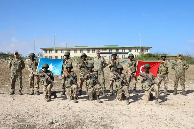 TÜRKSOM'da eğitim alan Somali askerlerinin fotoğrafı. Askerler ellerinde tüfekler ve üniformaları ile Türk ve Somali bayrakları tutarak poz veriyor.
