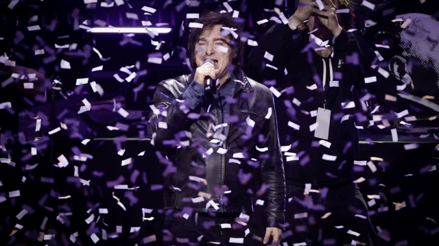 Javier Milei falando em um microfone durante evento. Há vários confetes caindo sobre ele