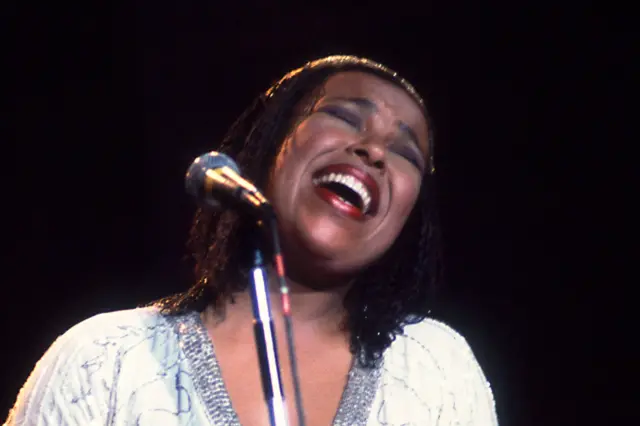 Roberta Flack is performing on stage with a microphone on a stand in front of he. She is wearing a dark outfit with a high neckline and has shoulder-length hair styled straight. The background is dark with a soft spotlight illuminating the figure, creating a dramatic concert atmosphere.