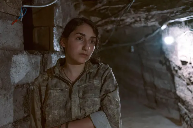 Portret Aryen w tunelu – ma na sobie mundur wojskowy, naszyjnik i bransoletkę. Jedną rękę ma skrzyżowaną na talii i tęsknie patrzy przed siebie. Światło na ścianie tunelu oświetla ją od tyłu.