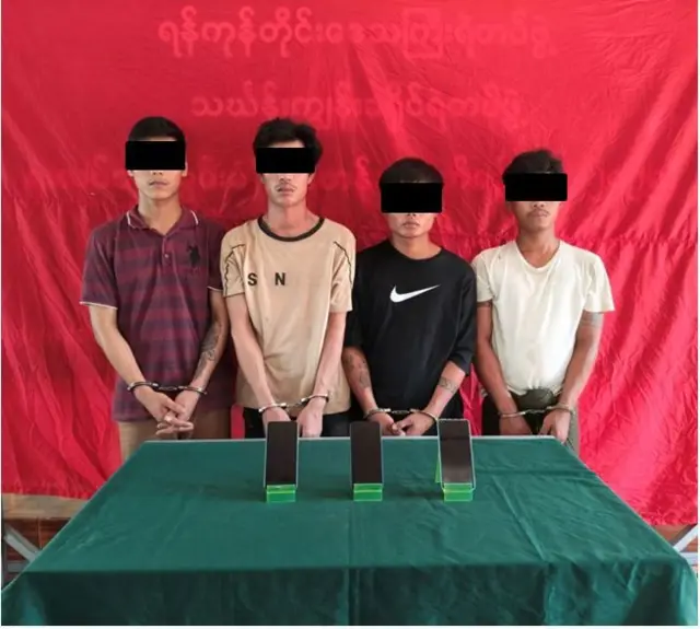 ဖုန်း ၃ လုံးတင်ထားတဲ့ စားပွဲအနောက်မှာ လက်ထိပ်ခတ်ခံထားရတဲ့ အမျိုးသား ၄ ဦး မတ်တပ်ရပ်နေပုံ 