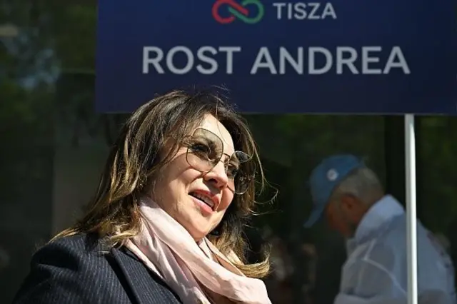 Rost güneş gözlükleriyle Rost Andrea yazılı bir bayrağın önünde gülümsüyor, pembe şalı ve gri kabanı var. 