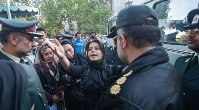 İranlı bir kadın, üniformalı iki ahlak polisiyle konuşurken eliyle işaret ediyor. Yanında başka kadınlar da var.