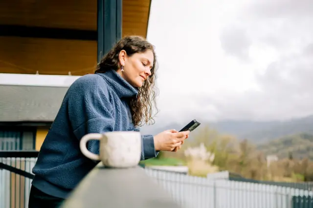 Mulher apreciando o café da manhã e usando o celular na varanda, com vista para a natureza.