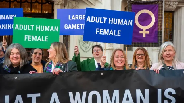 Women line up behind one black banner wey dem hold in front of dem. Some dey also hold signs above dia heads, including two wey read as "adult human female". 
