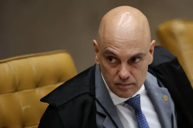 Moraes sentado um pouco inclinado para frente com semblante sério e a sombrancelha direita levantada, durante sessão no plenário do STF. Ele veste terno cinza, gravata azul e toga de juiz.