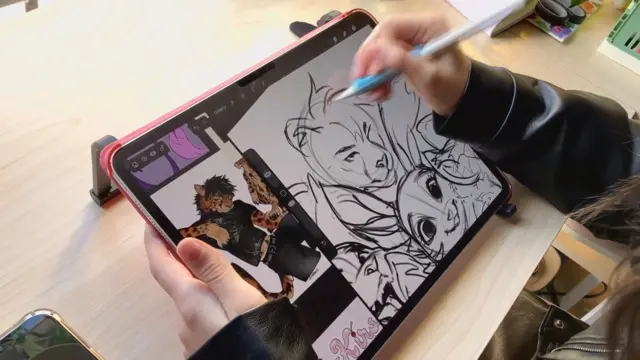 A tablet computer resting on a desk. It is being held by a person's left hand, while their right hand holds a white digital pencil, with which they're drawing on the screen. The image being drawn is a group of animals. They are sketched, line drawings. One of the creatures on the screen looks happy, while a big cat-type animal looks rather fierce. The person doing the drawing is wearing a black leather jacket. 