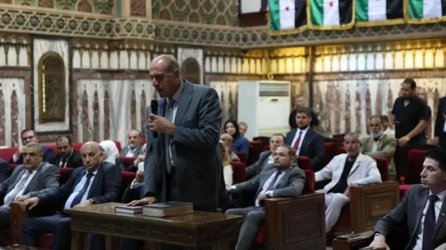 Yüksek Seçim Komisyonu'nun alt komitelerinin üyeleri 3 Eylül'de Şam'da yemin ederek göreve başlarken bir adam elinde mikrofonla, kutsal bir kitap olması muhtemel bir kitaba el basarak yemin ediyor