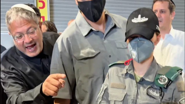 Bir adam elinin kaldırmı birilerini taciz eder gibi görünüyor, yanıda güvenlik görevlileri var.