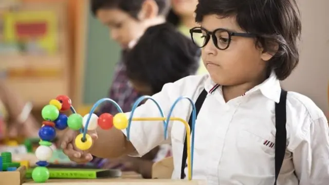طفل يرتدي نظارات طبية يلعب بلعبة تعليمية خشبية مكوّنة من خرز ملوّن وأسلاك معدنية داخل صف دراسي.