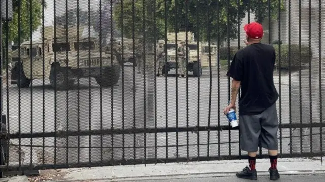 Мужчина перед решетчатыми воротам, за которыми военные машины