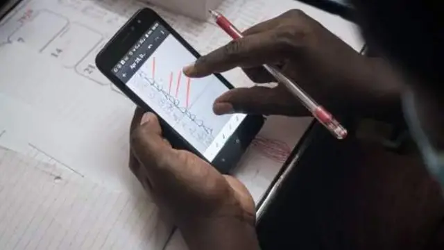 Wani mutum riƙe da waya ɗauke da hoton amsoshin jarrabawa a ciki, da kuma biro a ɗaya hannun nasa yayin da takardun jarrabawa ke kan teburinsa.