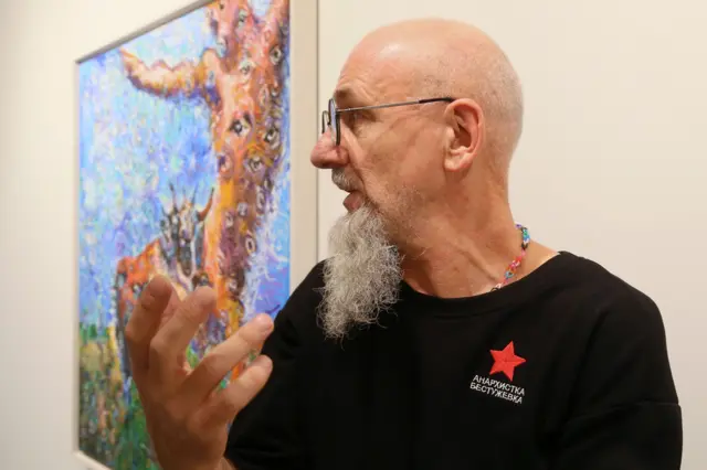 Мужчина с бритой головой и седой бородой, в черной футболке с красной звездой и в очках что-то рассказывает