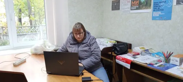 Жінка у фіолетовій куртці працює за коричневим столом з ноутбуком у пункті незламності 