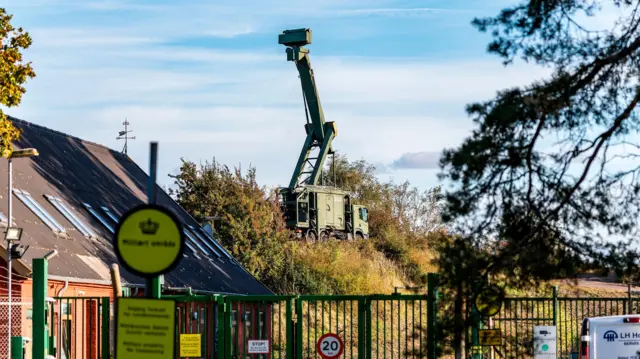 Радар установлен на верхушке крана, окрашенного в темно-зеленый военный цвет. На переднем плане видны кирпичные здания и зеленые ворота.