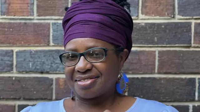Potret Angie Mkorongo di depan dinding bata. Ia mengenakan penutup kepala ungu, kacamata hitam, dan anting-anting melingkar bermanik-manik biru cerah, dan sedang tersenyum.