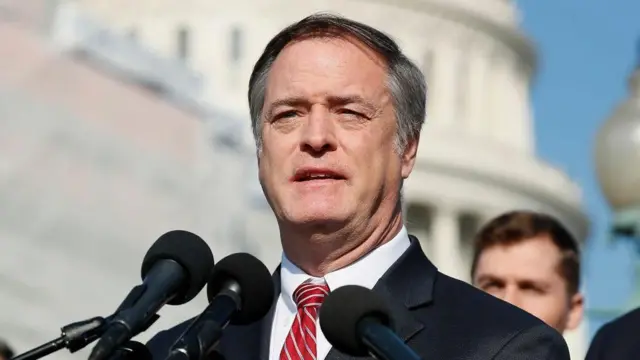 رابرټ بیلوټ د درېځ شا ته ولاړ دی او مخ ته یې درې مایکروفونونه دي. هغه خړ ویښتان لري. سپین کمیس او سره او سپینه نیکټای یې تړلې ده او توره کورتۍ یې اغوستې ده.