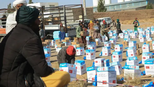 عکس نیمرخ از یک مرد افغان که منتظر کمکهای بشری است. پیشروی او بستههای از کمکها دیده میشود که برای هر فرد به گونه تقسیمشده روی زمین گذاشته شده است. دورتر چند فرد دیگر نیز دیده میشوند که بستههایی از این کمکها را بدست دارند.