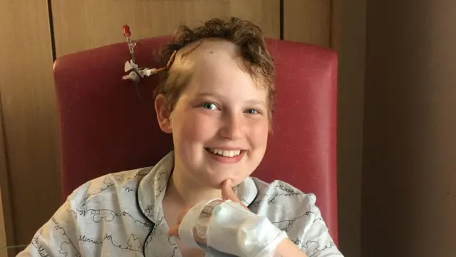 Una joven sonríe a la cámara. Está sentada en una silla roja y lleva pijama. Su mano izquierda está vendada y parece cubrir una cánula.