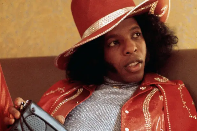 Sly Stone is dressed in a shiny silver top paired with a vibrant red leather jacket and a matching wide-brimmed hat. Both the jacket and hat are decorated with sparkling embellishments arranged in patterns. Sly is holding a gray rectangular object with a speaker grille and buttons, possibly a portable radio or electronic device. The background features a textured beige wall, suggesting an indoor setting.