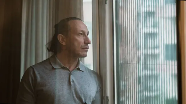 Çarkçı'nın camdan dışarı baktığı bir fotoğraf. Üzerinde gri bir kıyafet var. Uzun saçlarını arkadan bağlamış.