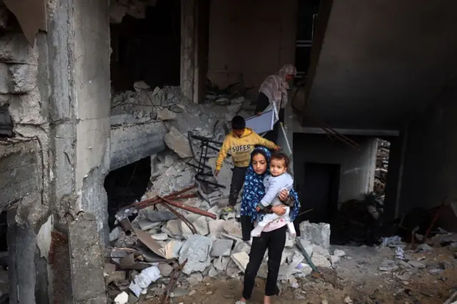 طفلة ترتدي حجاباً تحمل طفلة أخرى وخلفها طفل وامرأة يقفون على الأنقاض