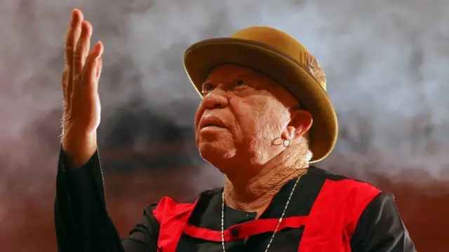 Salif Keïta en concert à Ankara, Turquie - 2022. Il porte un chapeau melon beige et un ensemble noir et rouge.