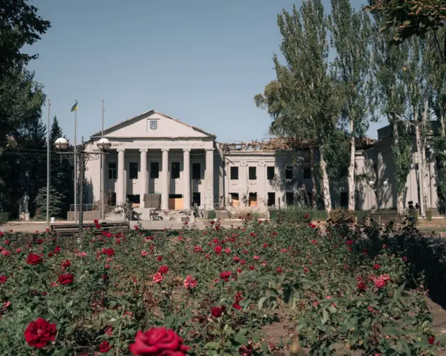 Велика будівля з колонами, яка частково зруйнована: видно вибиті вікна та пошкоджений дах. Перед нею – квітник із численними червоними трояндами. Будівлю оточують високі дерева, а небо чисте та ясне.