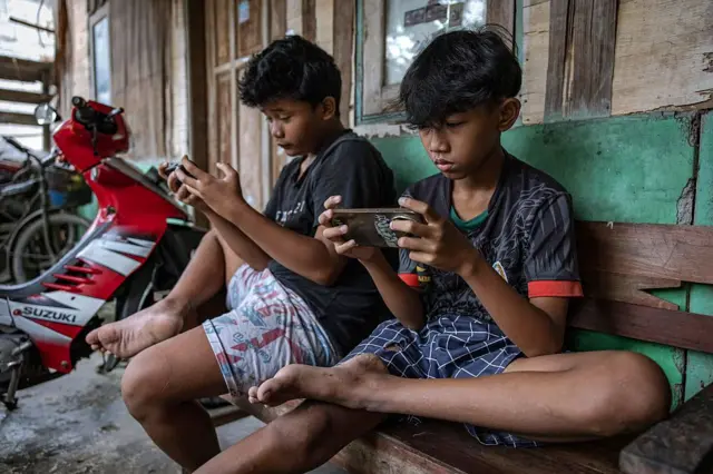 Dua anak laki-laki bermain game online di ponsel pintar mereka di depan sebuah rumah pada tanggal 27 Maret 2026 di Yogyakarta, Indonesia