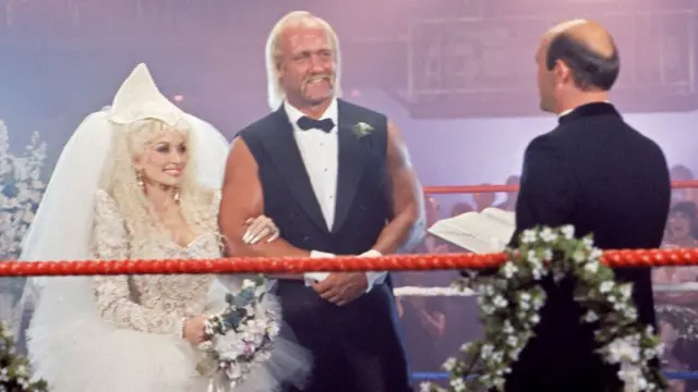 Hulk in a sleeveless suit and bow tie, arm in arm with Dolly Parton in a wedding dress in a wrestling ring, facing a priest holding an open Bible-style book on an episode of Dolly in 1987