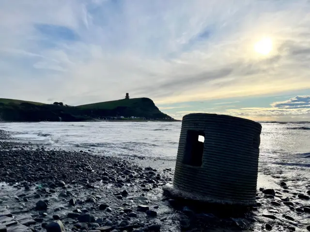 El fortín cilíndrico de hormigón se encuentra en una playa pedregosa. Al fondo se ven las colinas costeras.