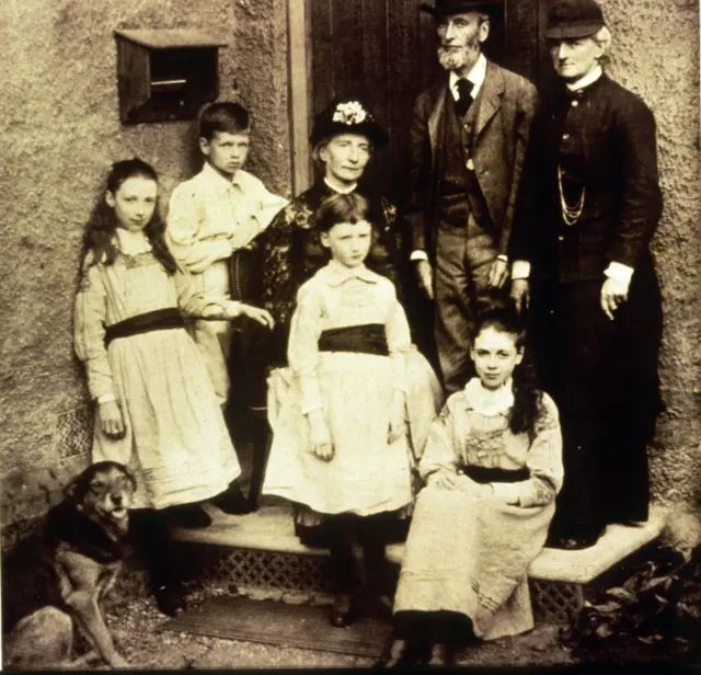 
Eglantyne sentada no degrau da entrada da casa com a tia e familiares.
