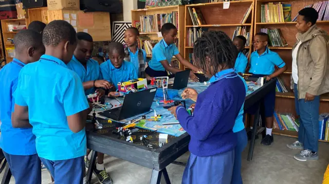 Abana benshi bambaye imyambaro y'ishuri y'ubururu. Bafite mudasobwa ku meza barimo no gukora za robo. Iruhande rwabo hahagaze umwarimu wabo w'umugore, wambaye ipantalo y'ikoboyi n'ikoti rya khaki. 