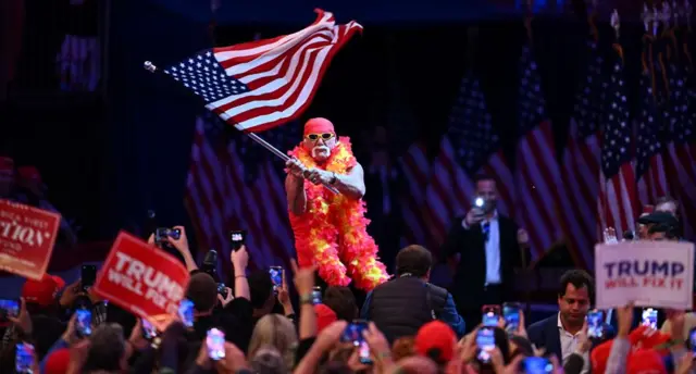Hulk on a stage waving a US flag in front of an audience, with some people holding Trump signs, at a campaign rally at Madison Square Garden in New York on October 27, 2024