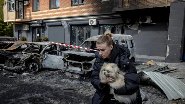 Жінка несе собаку біля зруйнованого будинку