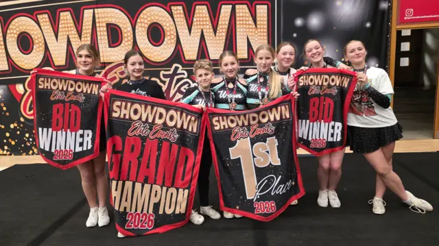 Ocho jóvenes se reunieron alrededor de varios carteles de los primeros clasificados. Llevaban mallas y estaban frente a una enorme marca. Participaban en una competición con mucha gente apiñándose detrás.