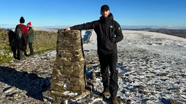 Un hombre se encuentra en la cima de una colina, con una ligera capa de nieve. Se apoya en un hito formado por un montón de piedras. Otras dos personas contemplan la amplia vista contra el cielo azul claro.