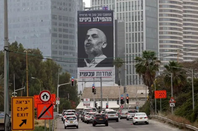 Carros em Tel Aviv passam por um outdoor com a foto de Sinwar, com uma mensagem em hebraico pedindo aos israelenses que se unam contra seu inimigo mais procurado