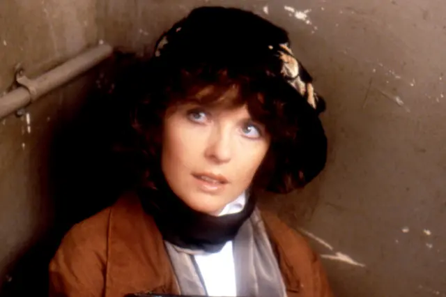 Diane Keaton photographed in costume during the filming of the 1980 film Reds. She is seated in a small, enclosed space with beige walls and a visible metal pipe running along the left side. She is wearing a brown coat over a white garment and has a dark hat adorned with a light-coloured patterned scarf or ribbon. The lighting is dim, creating a shadowy and confined atmosphere.