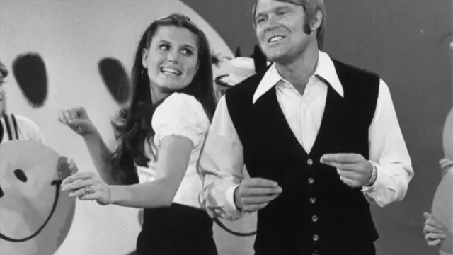Le chanteur Glen Campbell et l'actrice Lucie Arnaz, lors d'une émission de télévision américaine en 1971, avec des visages souriants en arrière-plan.