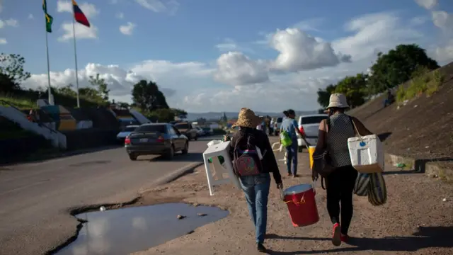 Imigrantes caminham carregando malas e bancos no acostamentouma estrada na fronteira entre Brasil e Venezuela