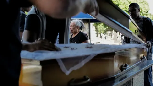 Divone é vista chorando ao fundo, com caixão da filha à frente